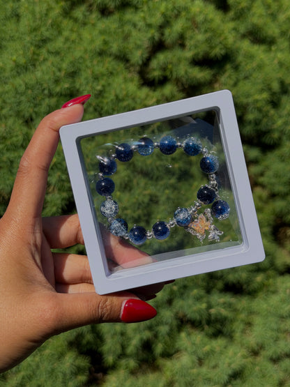 Blue beaded bracelet with silver accents and butterfly charm in clear display case, held in hand with red nails, green foliage background