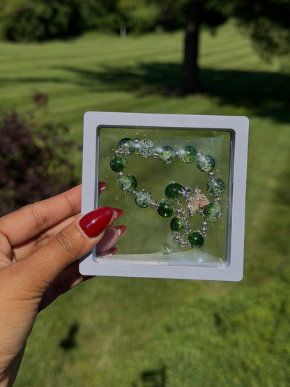 Green beaded bracelet with silver accents and iridescent charm in clear display case, held in hand with red nails, outdoor grass background