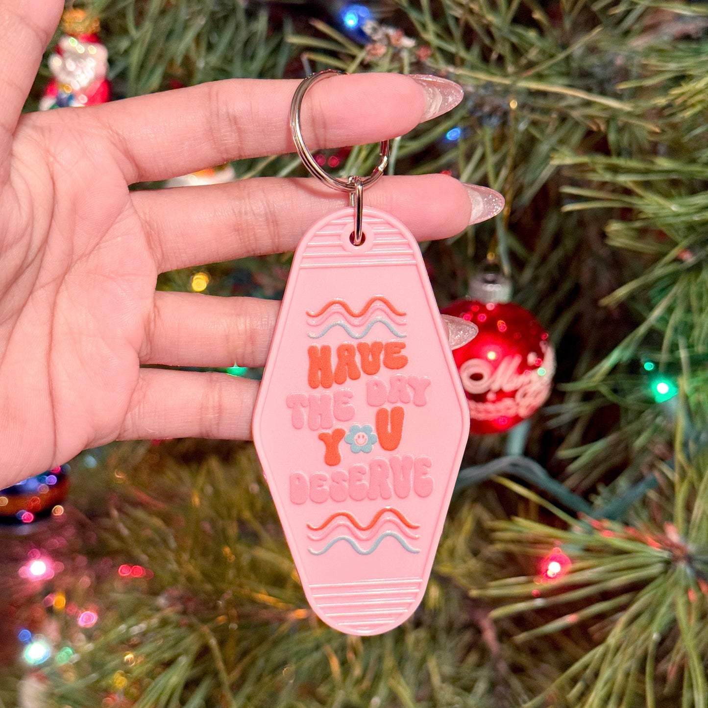 Pink motel keychain with "Have the day you deserve" text, against a Christmas tree backdrop.