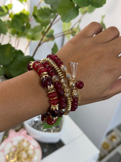 Layered beaded charm bracelets with various red, gold, and clear beads, adorned with gold-colored leaf and chain charms, on a tan wrist.