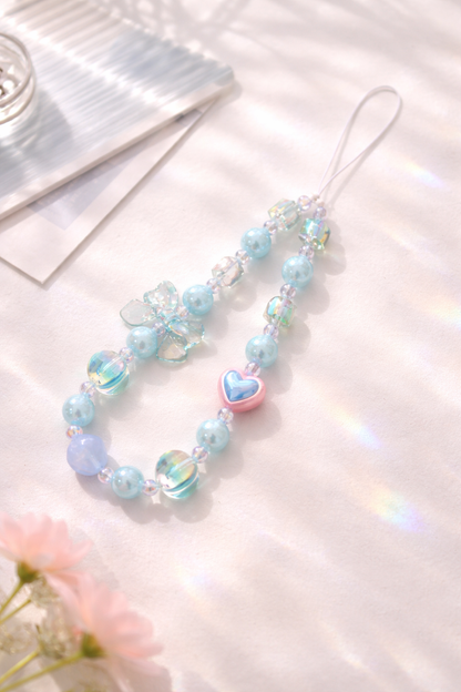 Beaded phone charm with light blue and iridescent beads, a pink and blue heart, and a clear butterfly, under subtle rainbow refractions.