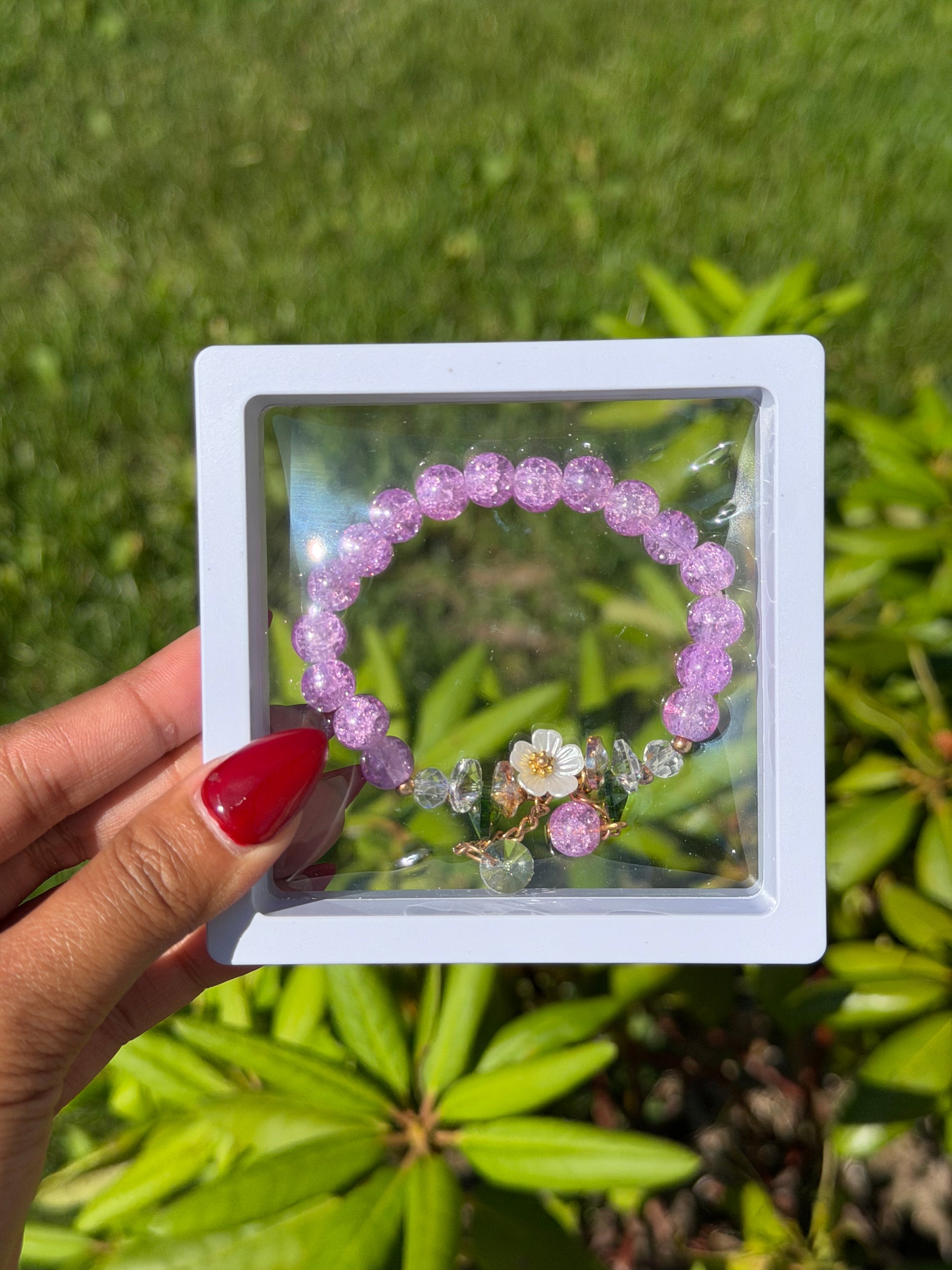 Purple beaded bracelet with clear and gold crystal charms and white flower accent in white display case, held against green foliage background