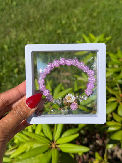 Purple beaded bracelet with clear and gold crystal charms and white flower accent in white display case, held against green foliage background