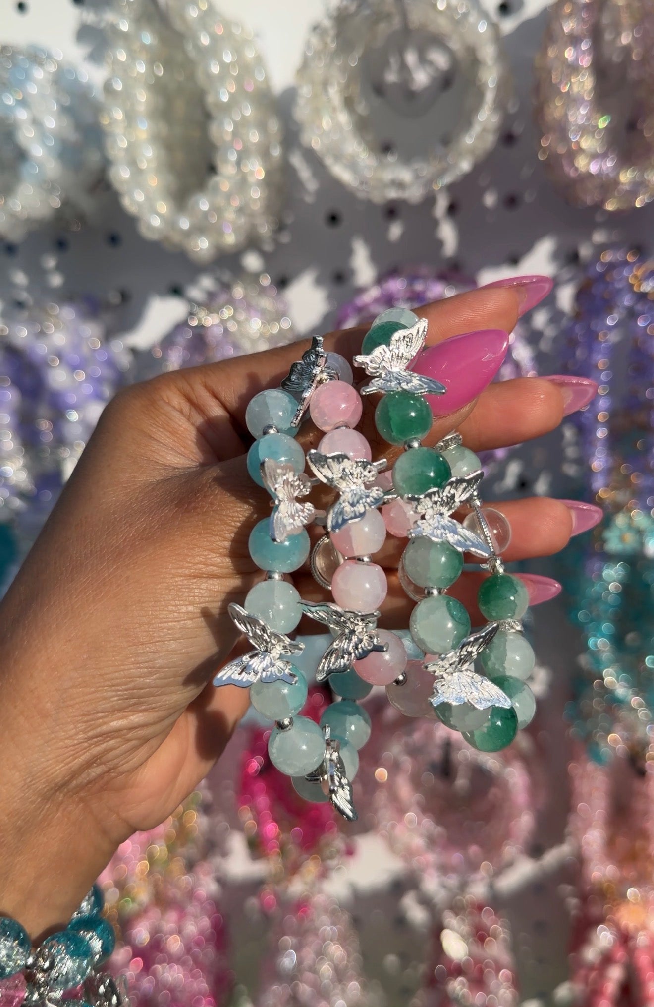 Beaded bracelet with light blue, pink, and green stones, adorned with silver butterfly charms, held in a hand with pink nails.