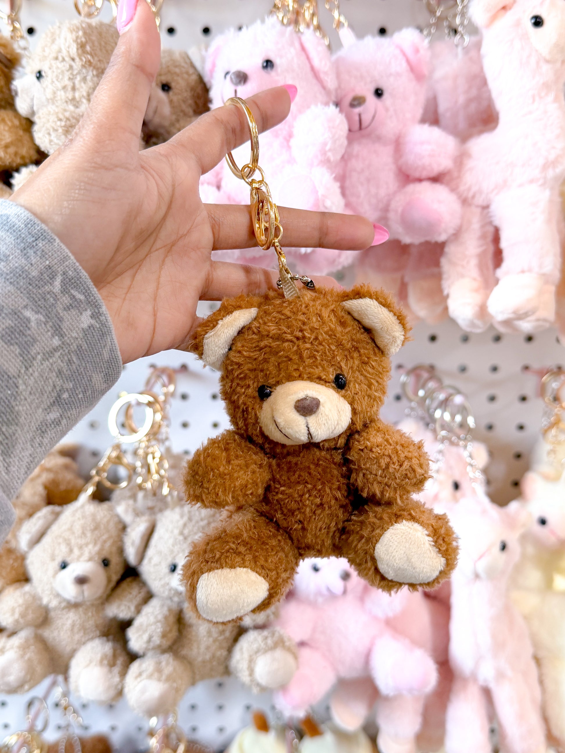 Brown plush Cuddle Bear Bag Charm with a gold keychain ring, held by a hand against a background of other teddy bear charms.