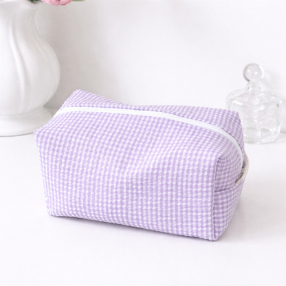 Pastel purple gingham makeup bag with a white zipper, on a white surface, alongside a white vase and glass decanter.