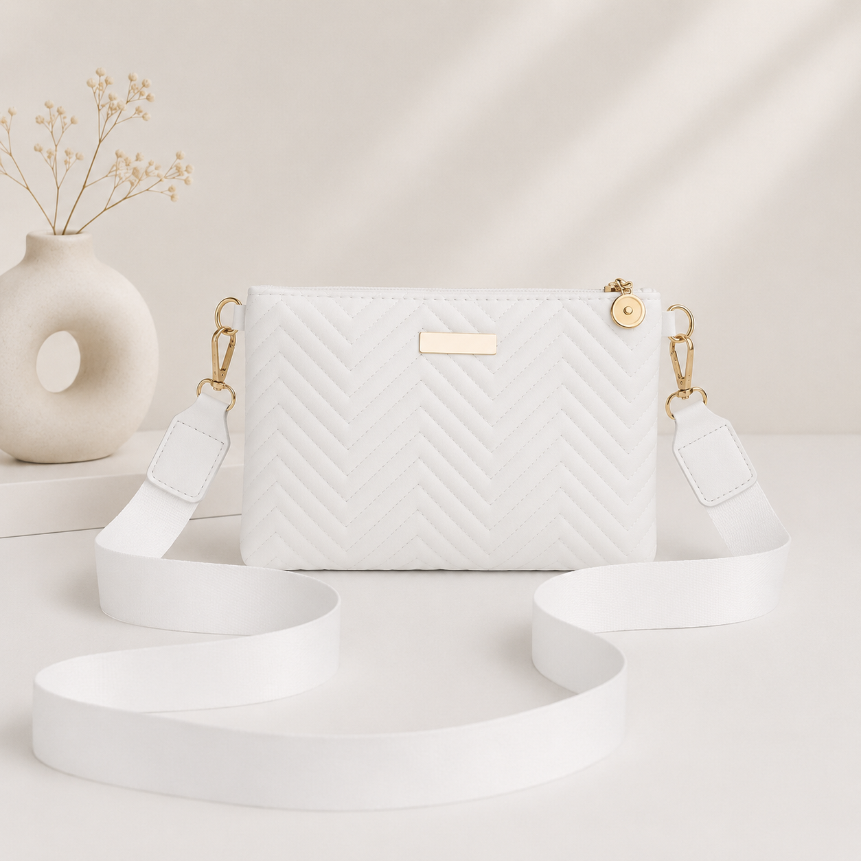 White quilted crossbody bag with wide strap and gold-tone hardware, front view on neutral background with decorative vase.