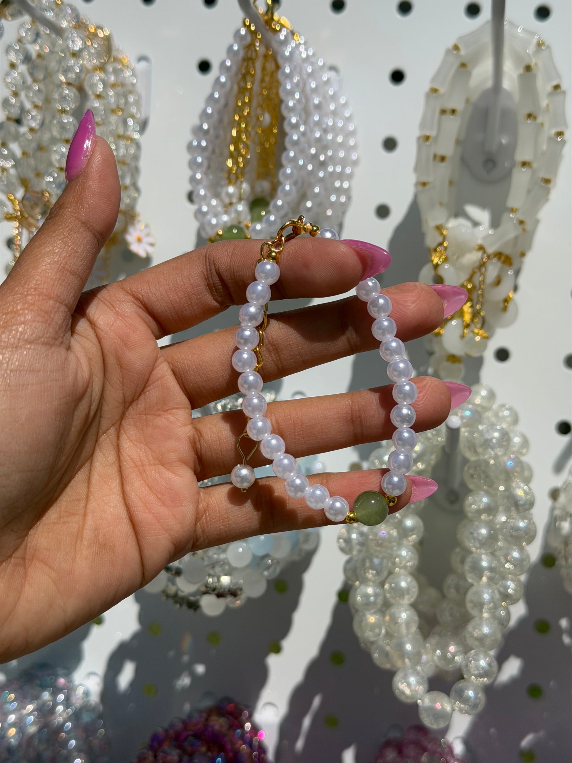 A white pearl beaded bracelet with a gold-tone clasp and a green bead, held by a hand with pink nails against blurred white bracelets.