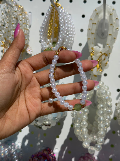 A white pearl beaded bracelet with a gold-tone clasp and a green bead, held by a hand with pink nails against blurred white bracelets.