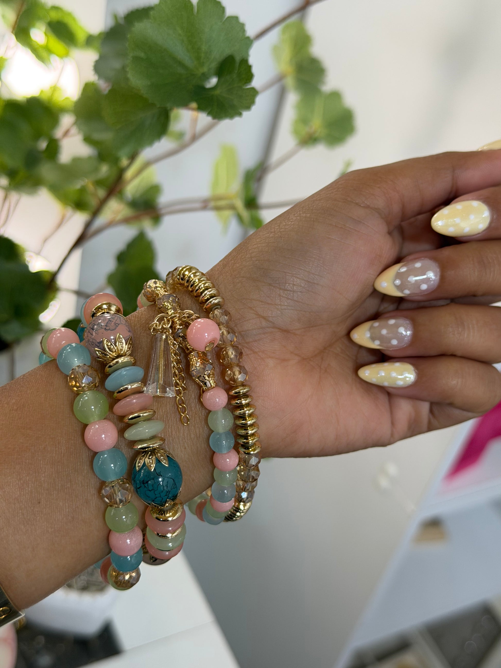 Multi-colored layered beaded charm bracelets with pastel pink, blue, and green beads, gold accents and charms, worn on a wrist with polka dot nails.