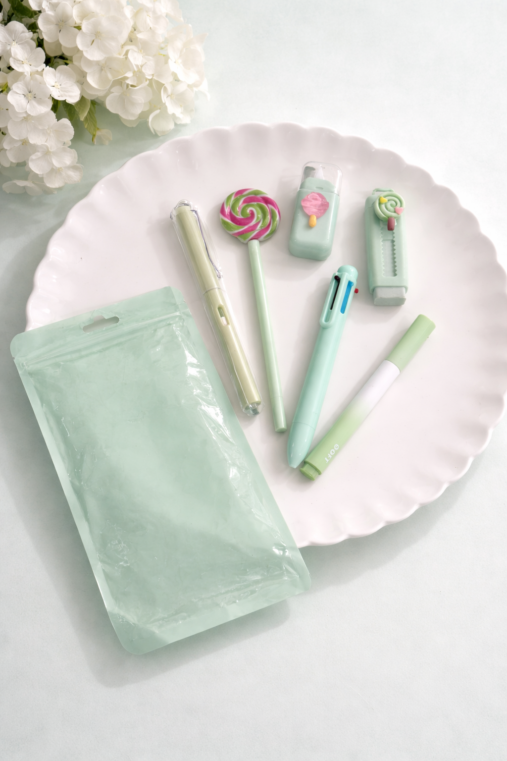 Mint green sweet desk set with pastel stationery on a white ruffled plate, next to a pale green pouch and white flowers.