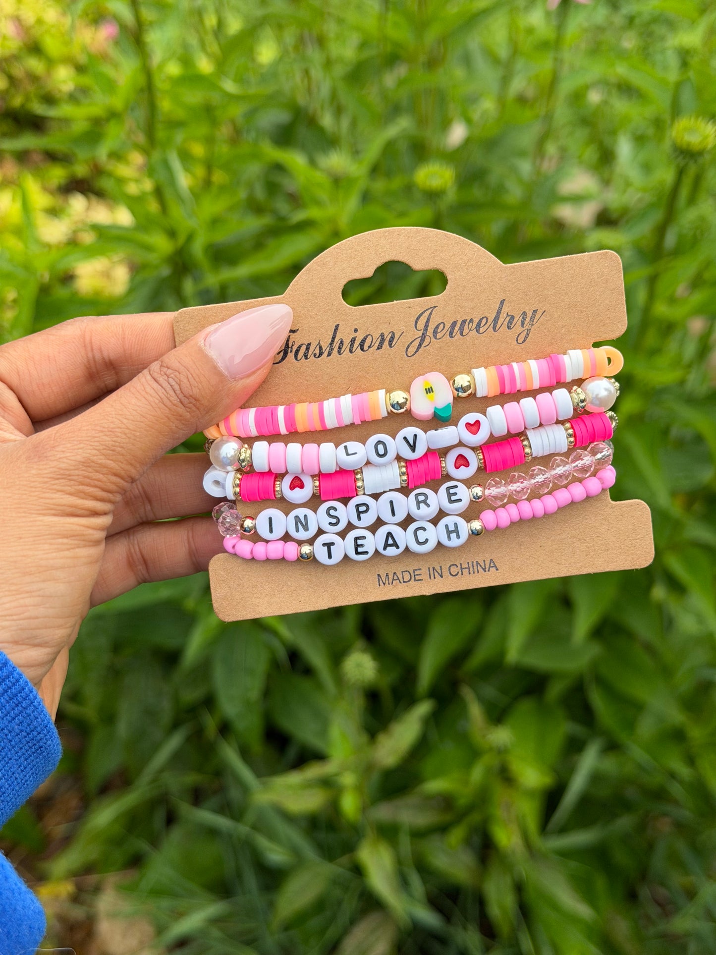 Colorful layered beaded bracelets with "LOVE," "INSPIRE," "TEACH" letter beads, pink and white accents, held on kraft card against green foliage.