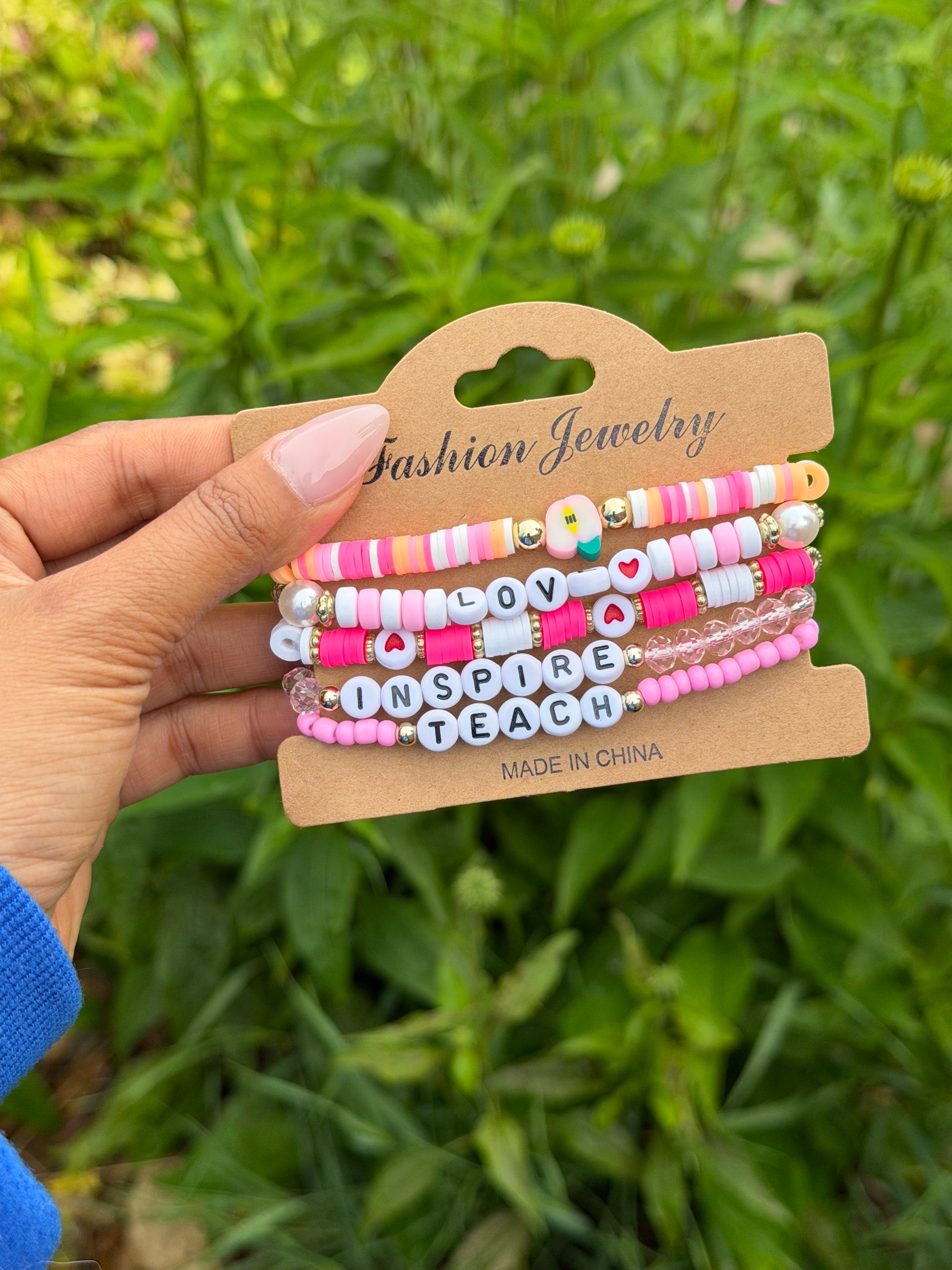 Colorful layered beaded bracelets with "LOVE," "INSPIRE," "TEACH" letter beads, pink and white accents, held on kraft card against green foliage.