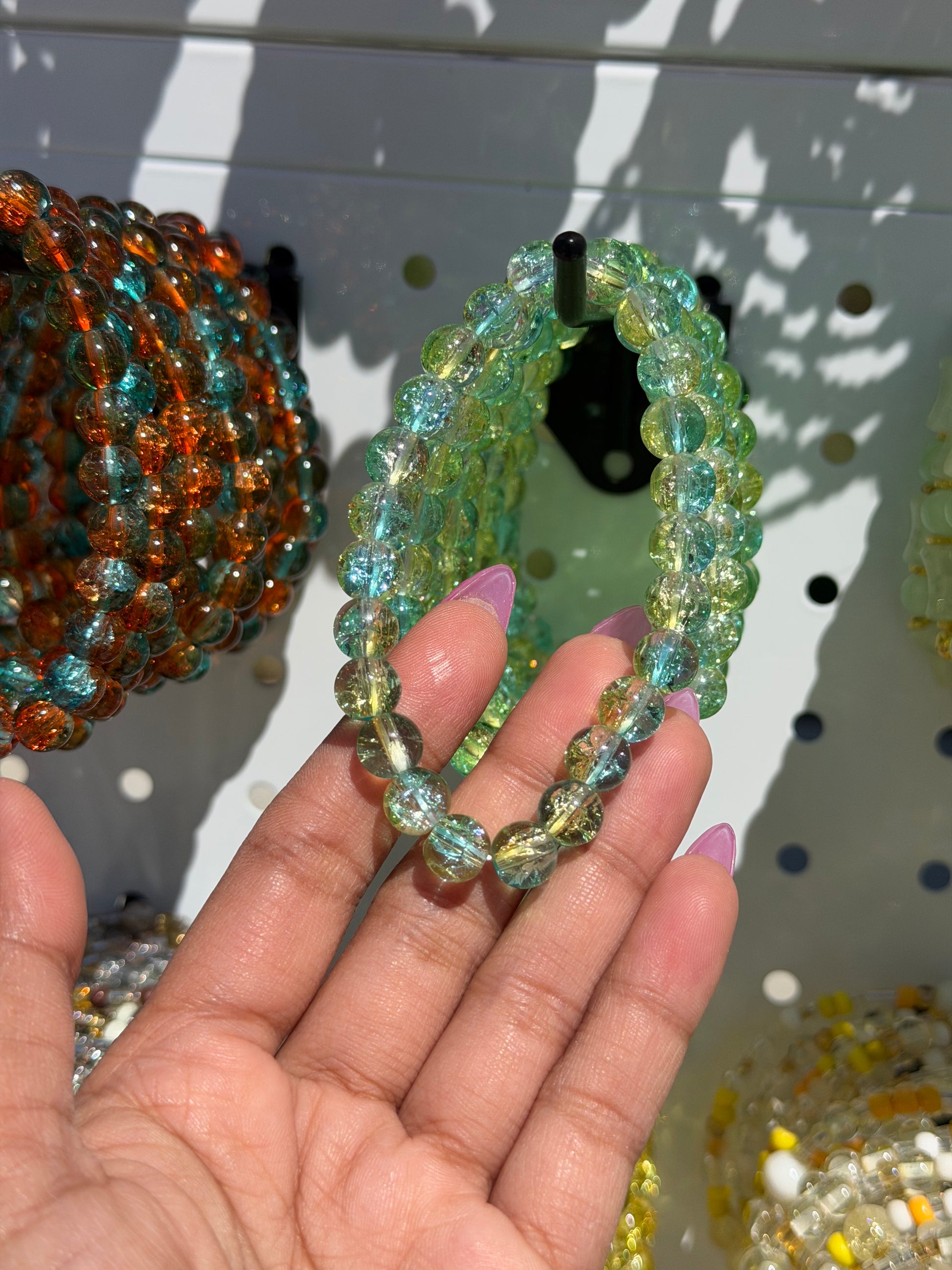 Green beaded bracelet with cracked glass beads in green and blue, held by a hand, with other colorful bracelets in the background.