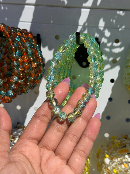 Green beaded bracelet with cracked glass beads in green and blue, held by a hand, with other colorful bracelets in the background.