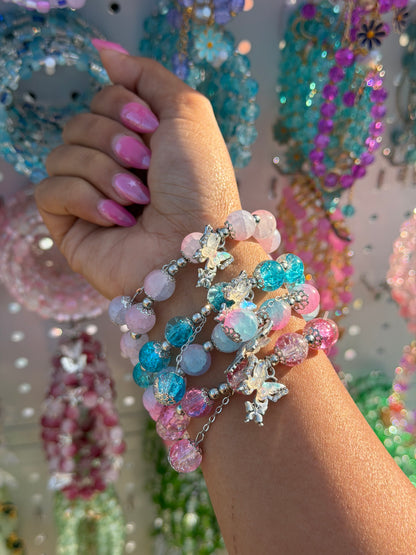 Monarch Charm Beaded Bracelet featuring pink, blue, and purple cracked beads with silver butterfly charms, stacked on a hand with pink nails.