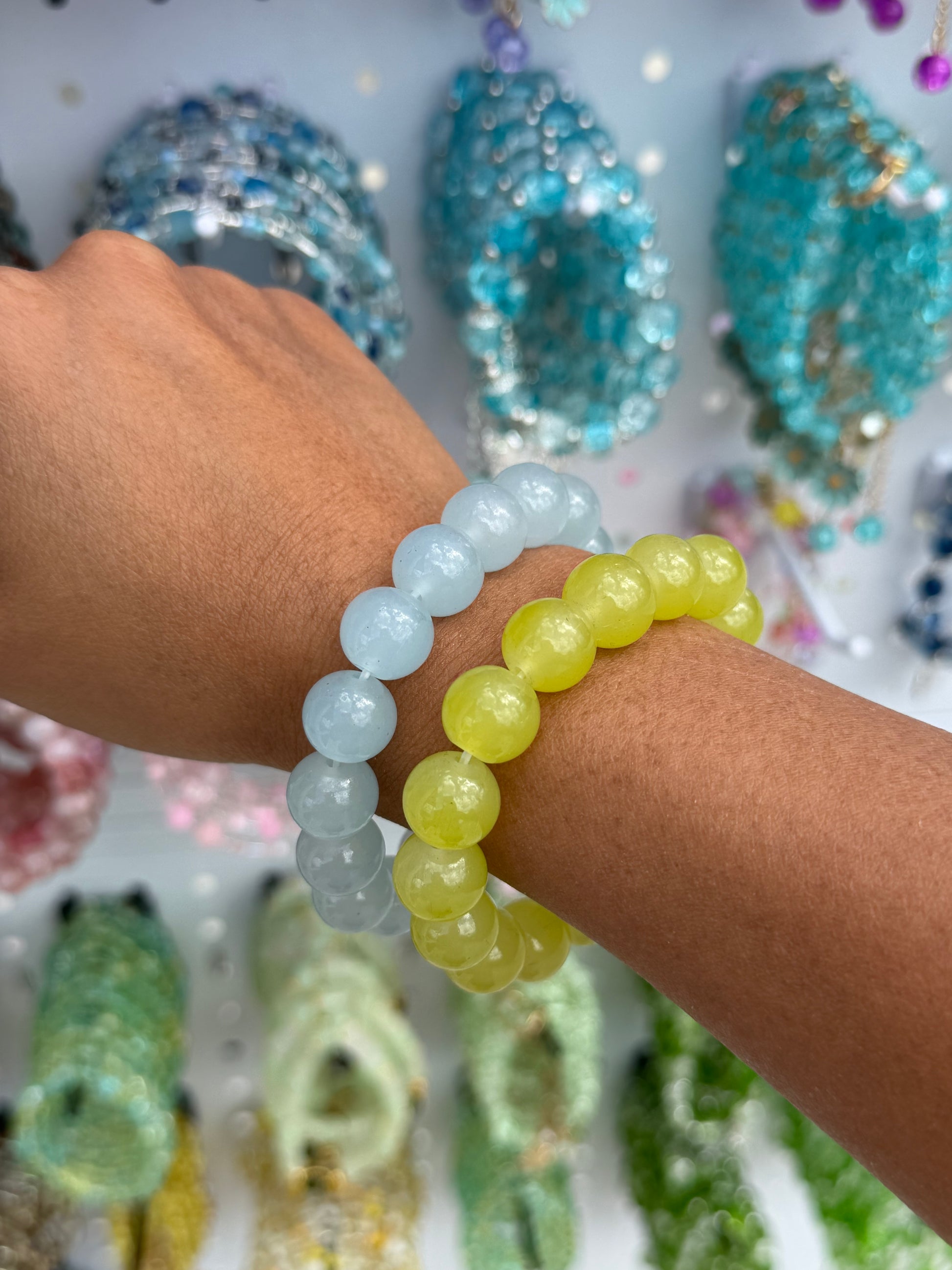 Pale blue and bright yellow beaded bracelets worn on a tanned arm, with blurred jewelry in the background.