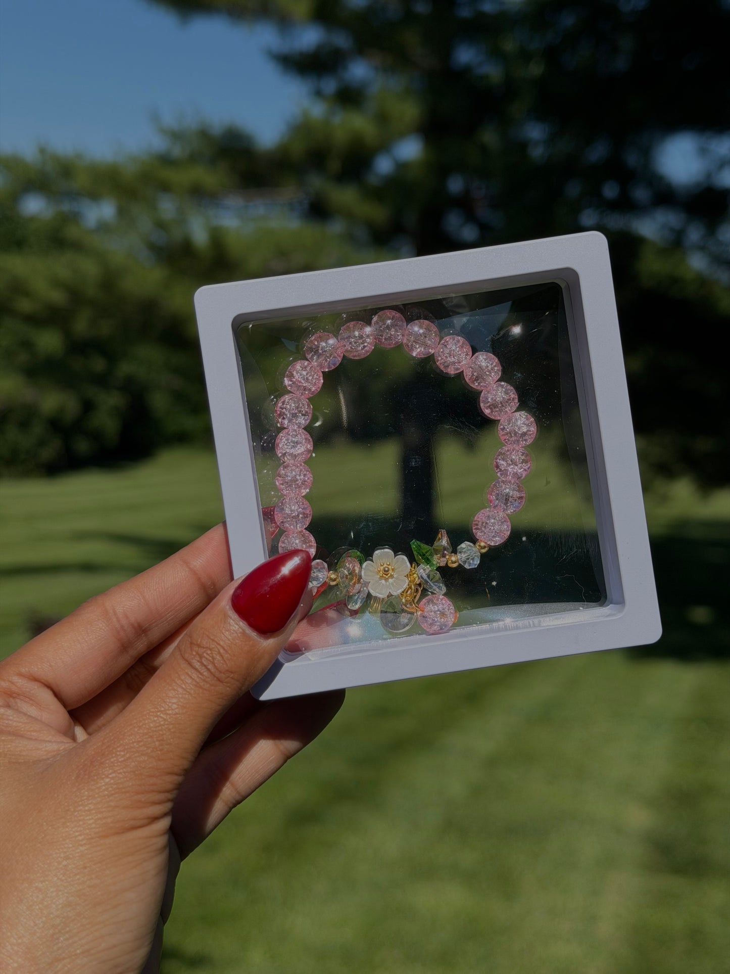 Pink beaded bracelet with floral charm and crystal accents in clear display case, held in hand outdoors