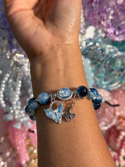 Blue butterfly charm beaded bracelet with iridescent and dark blue beads, featuring a silver butterfly and abstract cutout charm on a wrist.