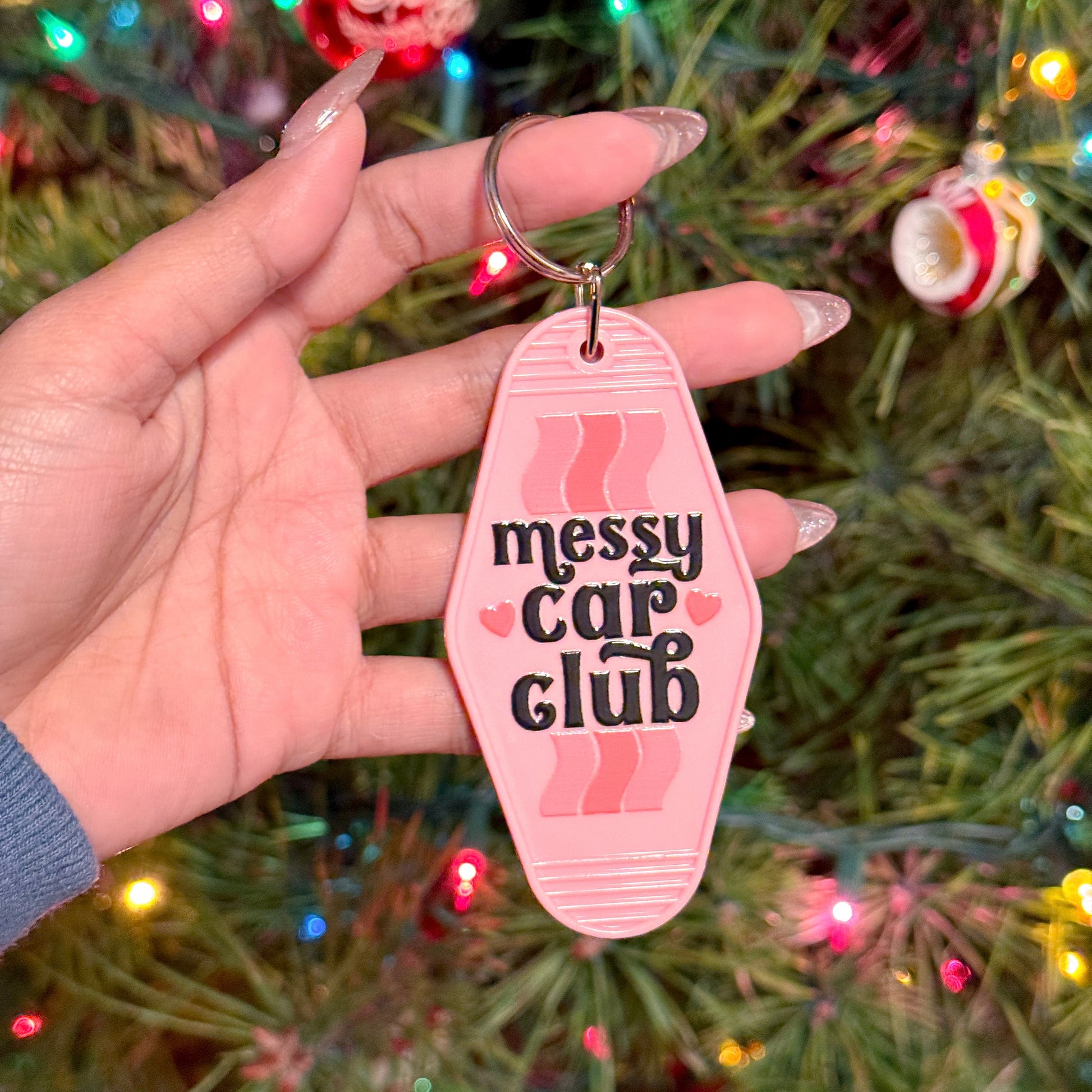 Pink motel keychain with "Messy Car Club" by an unspecified brand, displayed against a Christmas tree.
