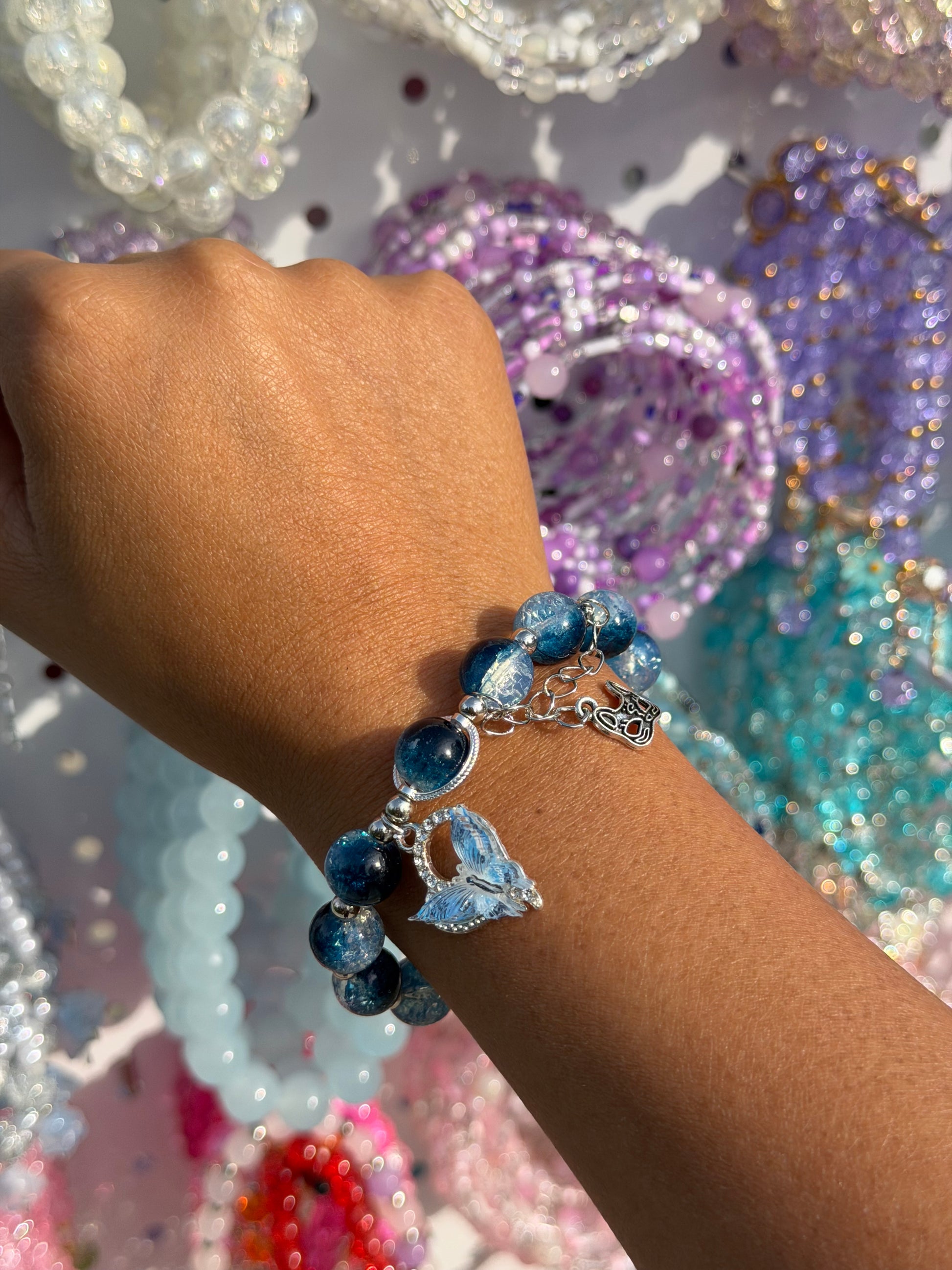 Blue beaded bracelet with a butterfly charm worn on a tanned arm, surrounded by blurred colorful bracelets.