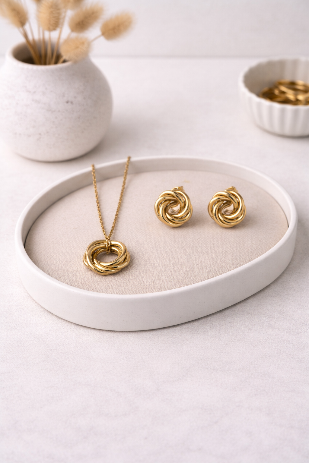 Gold knot necklace and matching stud earrings set displayed on a light beige cushion in a white oval tray.