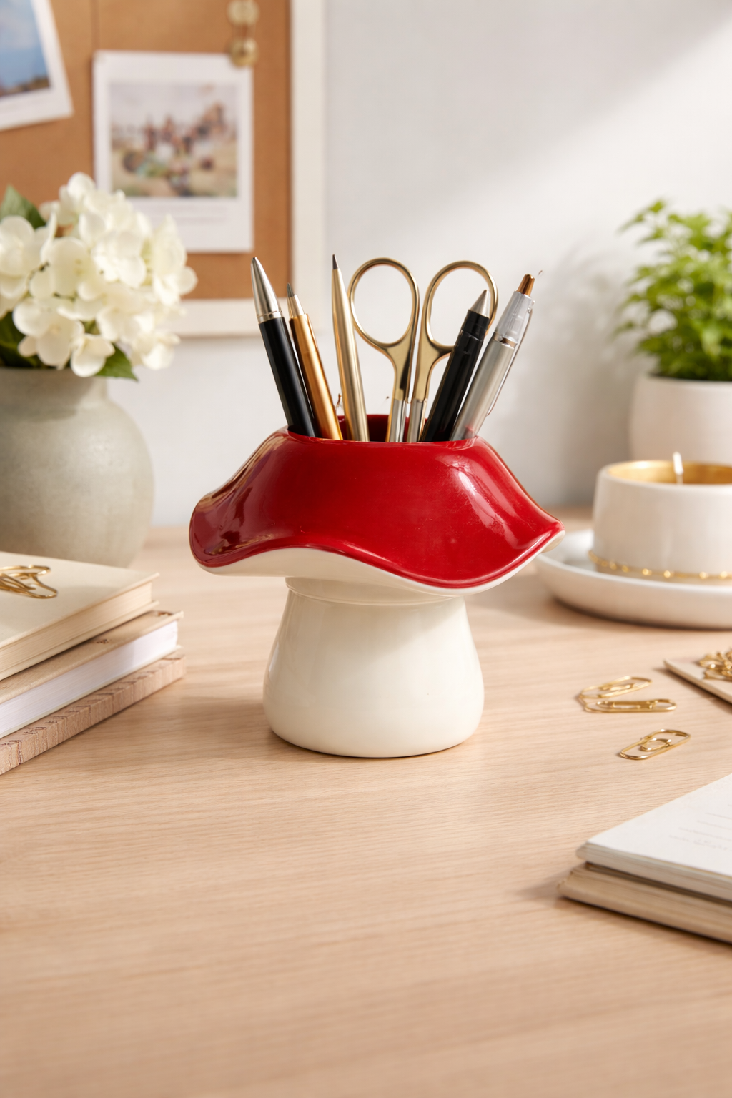 Glossy red cap and white stem mushroom pen holder on a light wood desk, filled with pens and scissors.