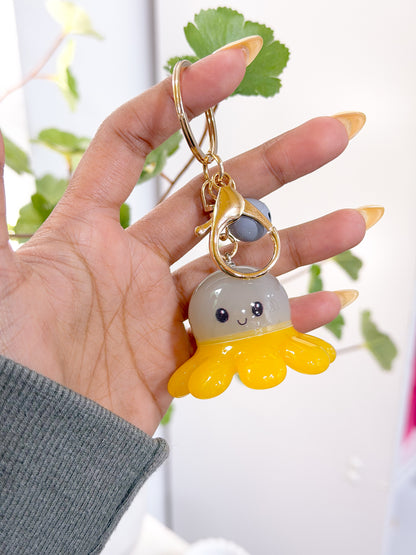 Glow Baby Octopus Charm with a grey head, yellow tentacles, and a smiling face, attached to a golden keychain, held in a hand.