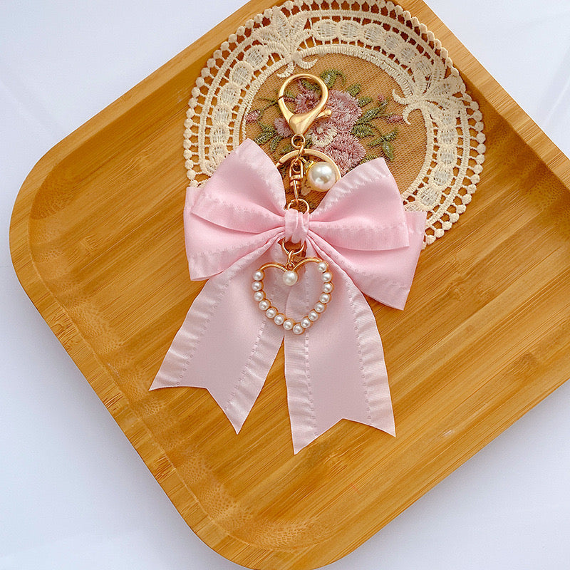 Pink satin bow pearl bag charm with a gold clasp, dangling pearl, and pearl-lined heart, on a wooden tray with a lace doily.