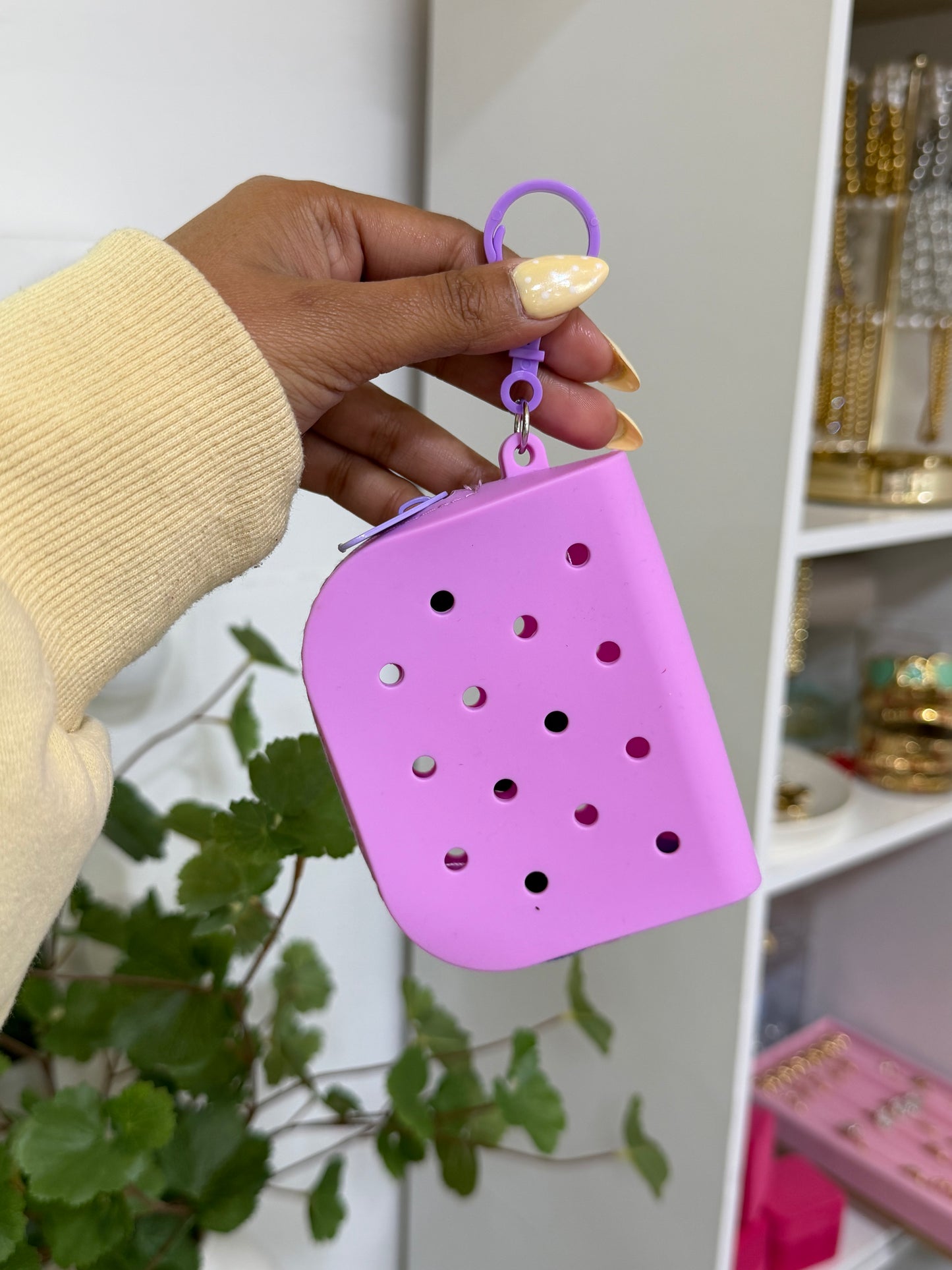 Lilac silicone charm pouch with croc-style holes, a zipper, and a purple carabiner, held by a hand with polka dot nails.